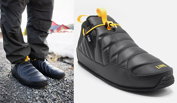 Person standing on gravel wearing Line Apres Booties slippers. Slippers have a thick cushioned with black outer and yellow detail.